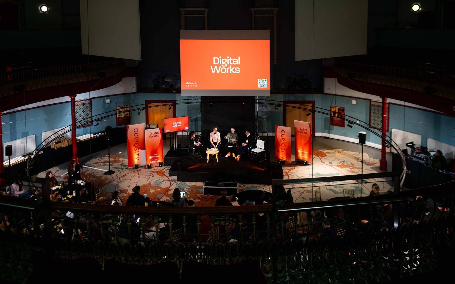 A photo of The Broderick Hall at Leeds City Museum taken from the balcony in the room, there is a large screen with an orange 'Digital Works' graphic on it, and 4 speakers sat in a row on brightly lit stage. The rest of the room is in darkness